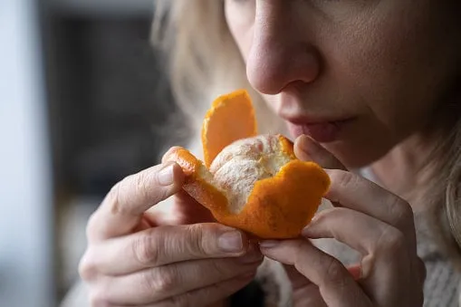 sick woman trying to sense smell of fresh tangerine orange, has symptoms of covid-19, corona virus infection - loss of smell and taste, standing at home one of the main signs of the disease