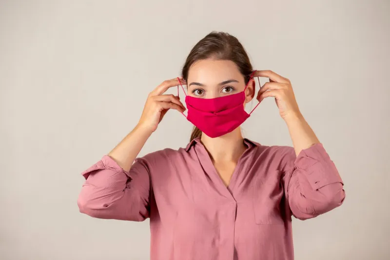 youthful woman concerned about coronavirus putting on a red cloth mask to protect herself female nurse in the process of attaching a medical mask