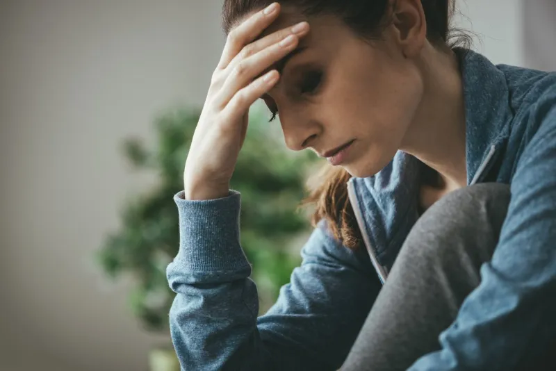 sad depressed woman at home sitting on the couch, looking down and touching her forehead, loneliness and pain concept