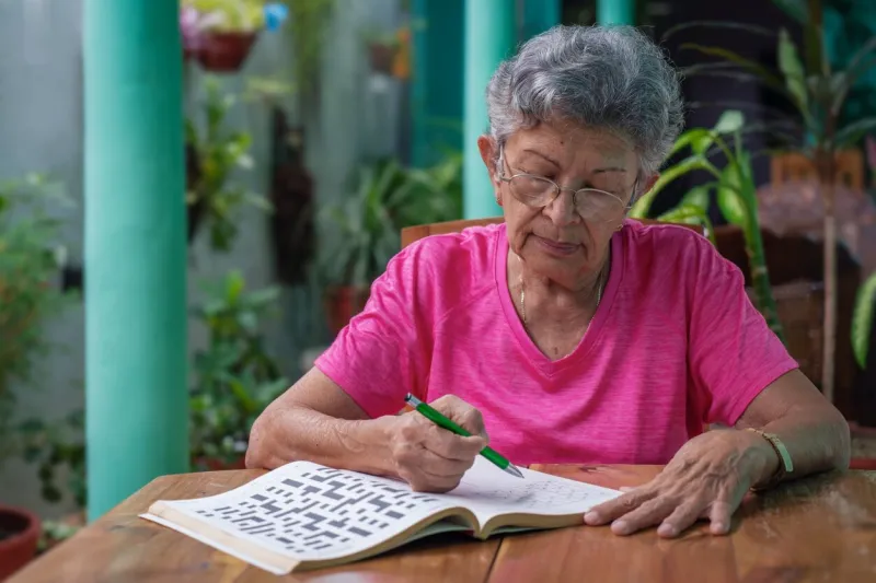 senior woman with glasses sitting at a table, filling in a sudoku puzzle