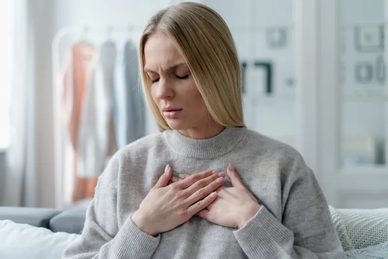 stressed adult female with blonde hair feeling sick at home apartment and touching heart area, cardiac pain symptom