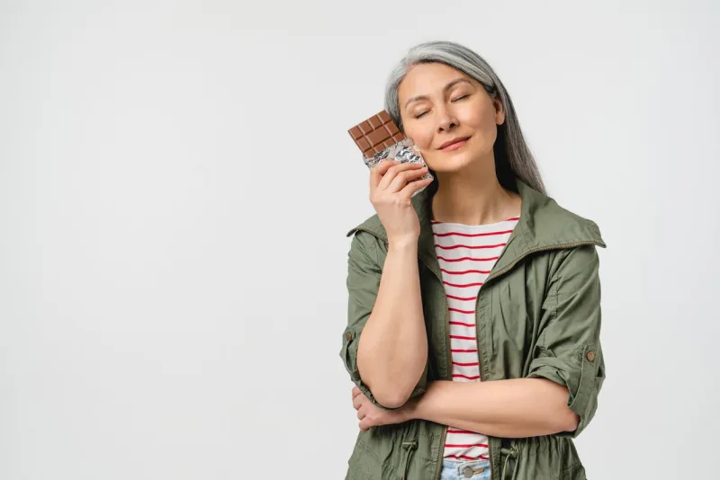 dreamy caucasian mature middle-aged woman with grey hair in casual clothes wants to eat chocolate bar full of calories isolated in white eating disorder concept junk fat unhealthy food dieting