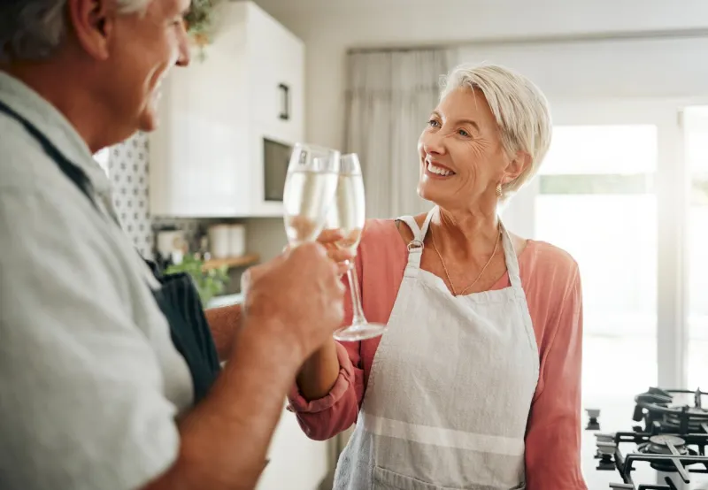 senior couple, champagne and drinks toast in house or home kitchen in marriage anniversary, celebration event or retirement security success happy smile, love or elderly man, woman and alcohol glass