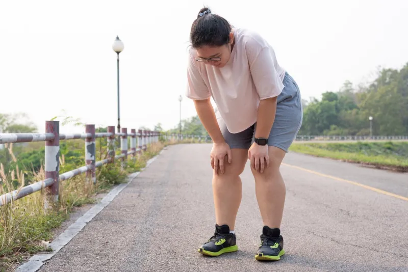wide shot portrait of overweight asian woman wearing eyeglasses, standing outdoor, bending down, resting after running, being tired, suffering from knee and joint injury sport and obesity concept