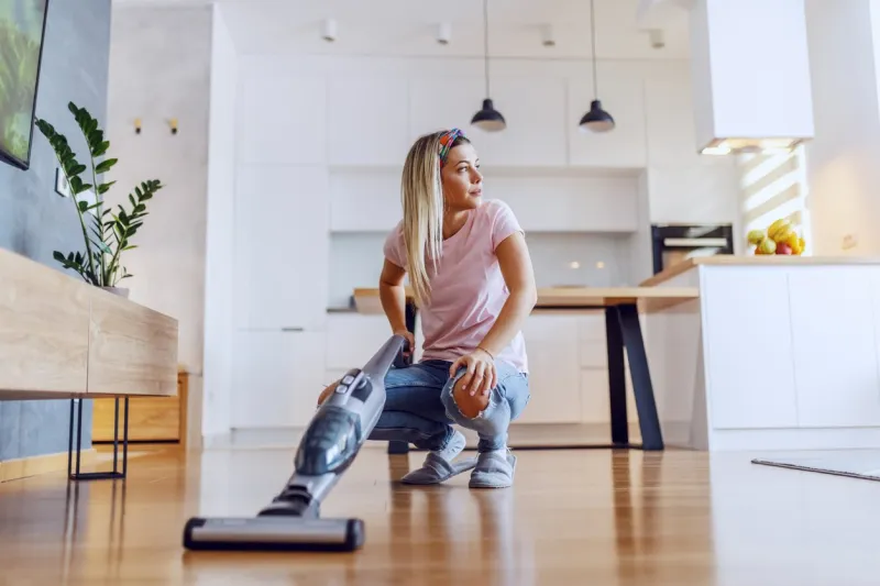 worthy caucasian blond housewife crouching in living room and using steam cleaner to clean parquet