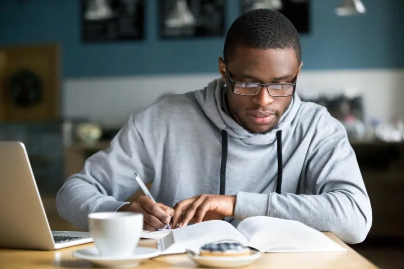 focused millennial african american student in glasses making notes writing down information from book in cafe preparing for test or exam, young serious black man studying or working in coffee house