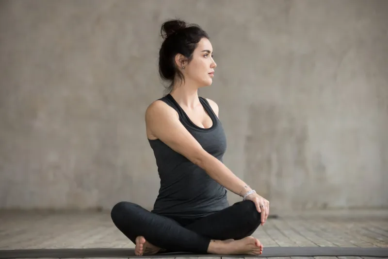 young sporty woman practicing yoga, doing revolved easy exercise, parivrtta sukhasana pose, working out, wearing sportswear, black pants and top, indoor full length, gray wall, yoga studio