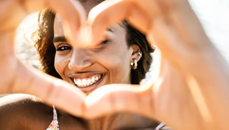 close up image of smiling woman in swimwear on the beach making a heart shape with hands - pretty joyful hispanic woman laughing at camera outside - healthy lifestyle, self love and body care concept
