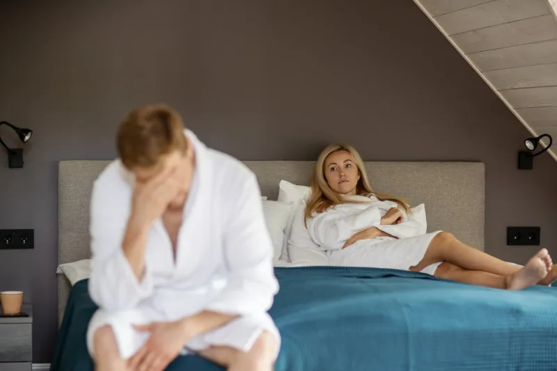 angry dissatisfied woman in white bathrobe lying on bed while her defocused depressed husband or lover rubbing forehead in foreground