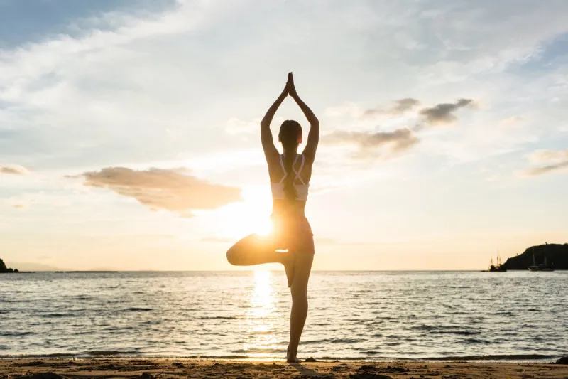 full length rear view of the silhouette of a woman standing on one leg while practicing the tree yoga pose on a tranquil beach, shot at sunset during summer vacation in indonesia