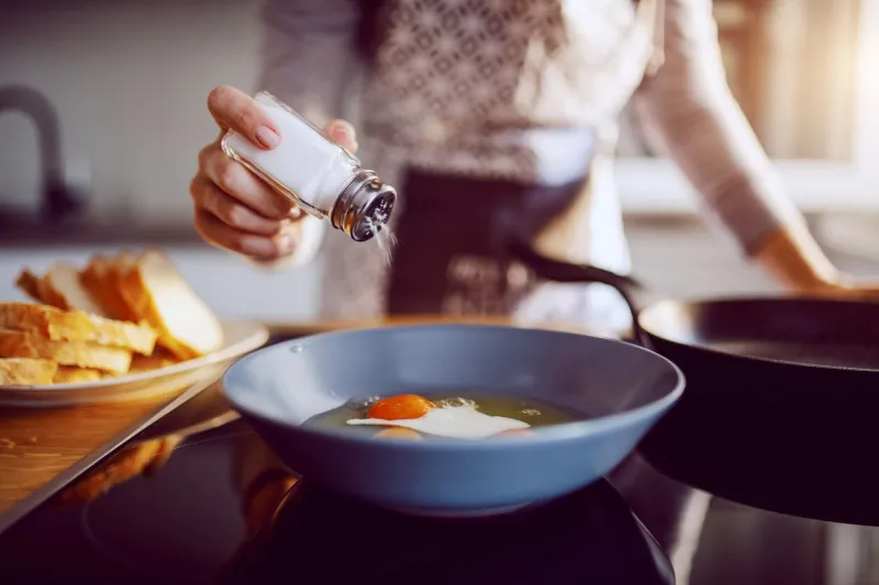 close up of caucasian woman adding salt in sunny side up eggs while standing in kitchen next to stove