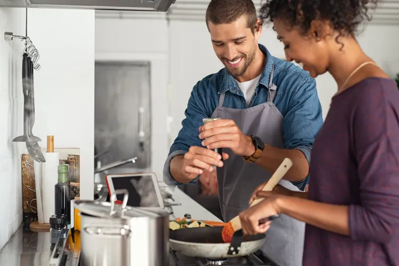 young man cooking with girlfriend and adding spice to the sauce guy adds black pepper into frying pan on stove while woman using spatula to mix multiethnic couple preparing lunch together at home