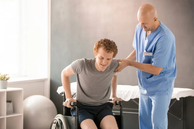 mature clinician in uniform helping young man in pain to sit in wheelchair after rehabilitation training while supporting his arm and elbow