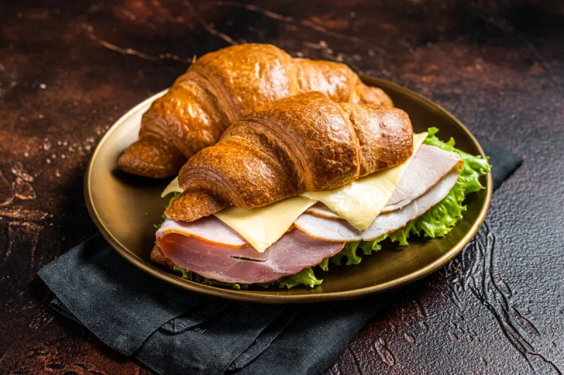 french croissant sandwich with lettuce, cheese, tomatoes and ham dark background top view