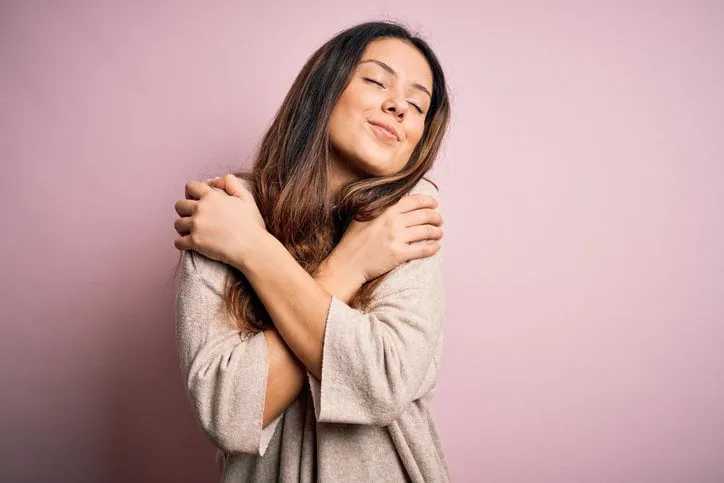 young beautiful brunette woman wearing casual sweater standing over pink background hugging oneself happy and positive, smiling confident self love and self care