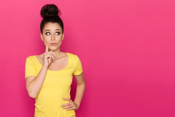 beautiful young woman in yellow top is holding hand on chin, looking away and thinking waist up studio shot on pink background