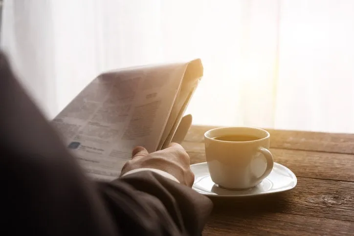 a man in a business suit in the morning in the sun reads the news in the newspaper, next to a cup of black coffee reading business news place for text