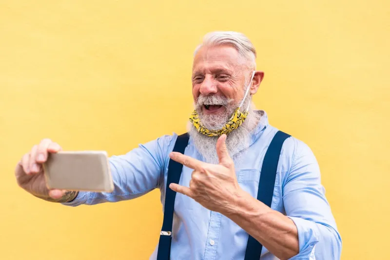 trendy hipster man using mobile phone doing video call while wearing fashion silk safety mask - outfit, technology and lifestyle during coronavirus time - focus on face