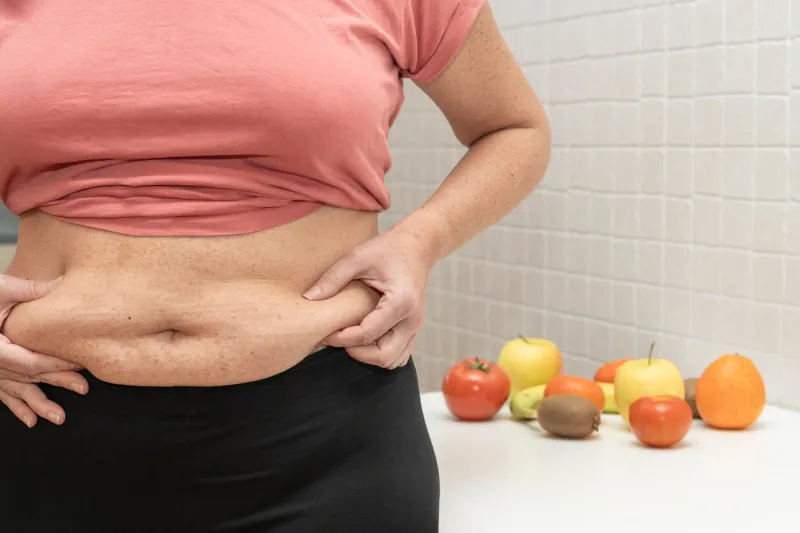 middle-aged woman shows excess abdominal fat and in the background various fruits and vegetables concept of weight loss by eating healthy