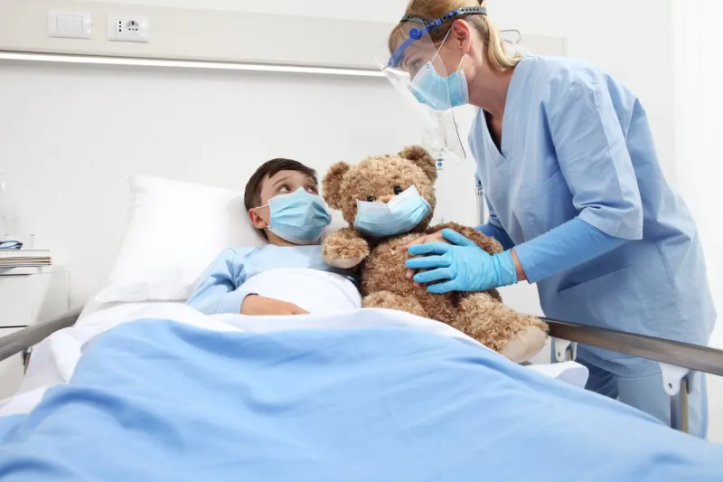 nurse takes care of the patient child in hospital bed playing with teddy bear, wearing protective masks, corona virus covid 19 protection concept
