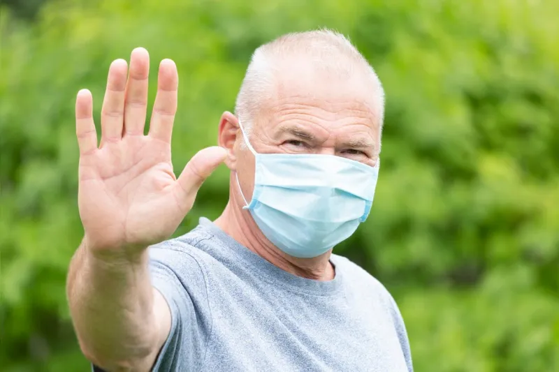 an elderly man with a protective mask makes a stop gesture with his hand looking directly at the camera against the background of nature