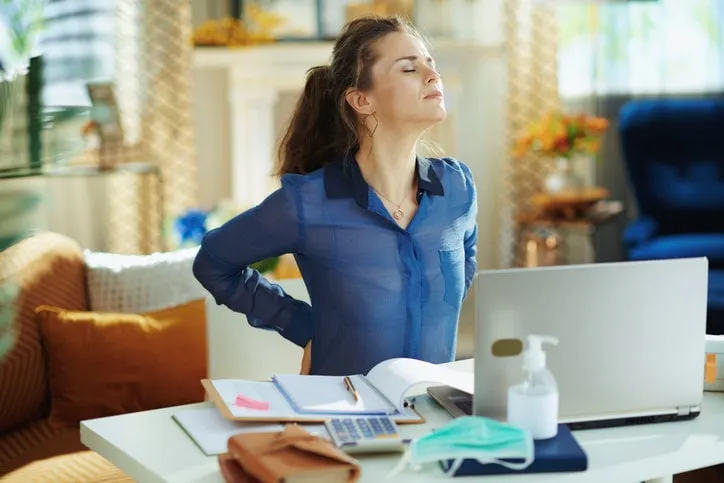 tired modern woman in blue blouse in the modern house in sunny day having back pain in temporary home office during the coronavirus epidemic
