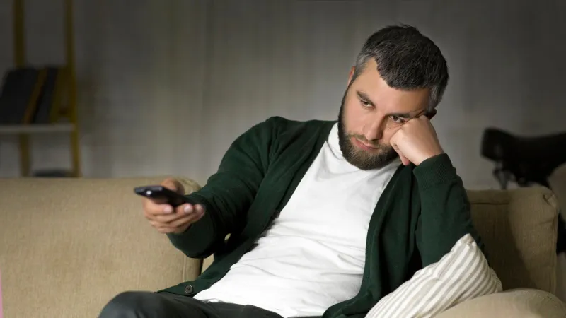 a man is casually lounging on a couch, holding a remote control with a bored look on his face while watching television the setting is a cozy living room illuminated by soft indoor lighting