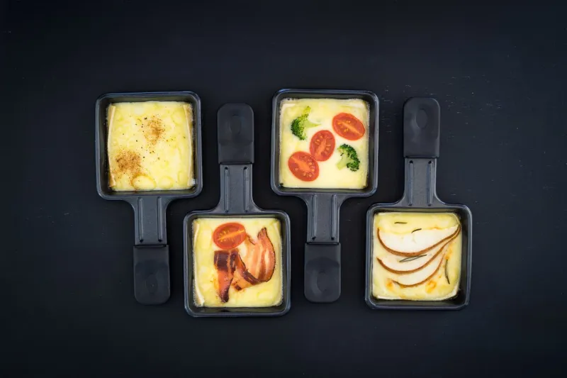four small square raclette pans with melted cheese and different ingredients  paprika, bacon, cherry tomatoes, broccoli and pear slices