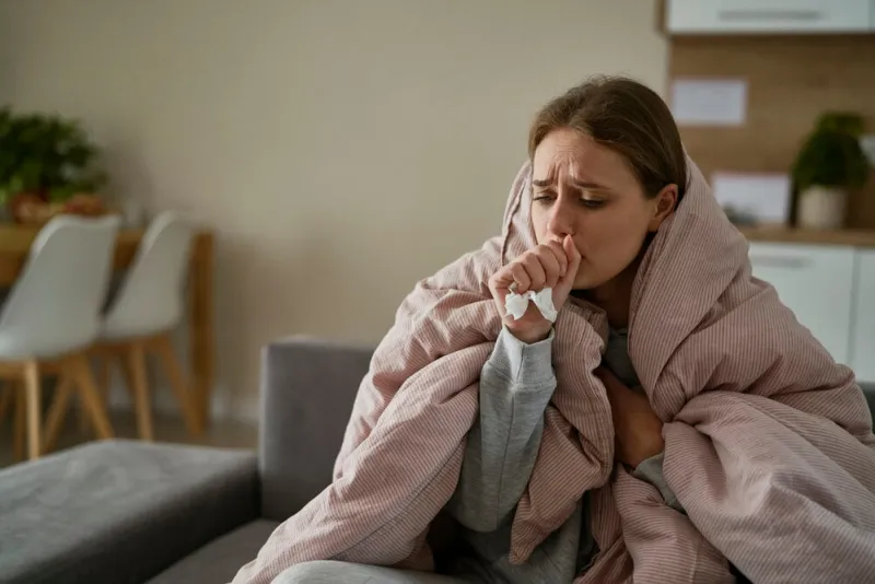 caucasian young woman coughing and sitting under the duvet at home