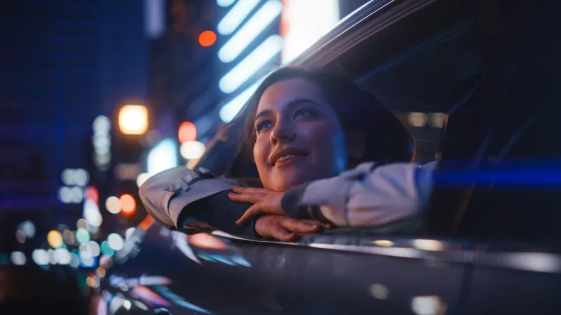 excited young female is sitting on backseat of a car, commuting home at night looking out of the window with amazement of how beautiful is the city street with working neon signs
