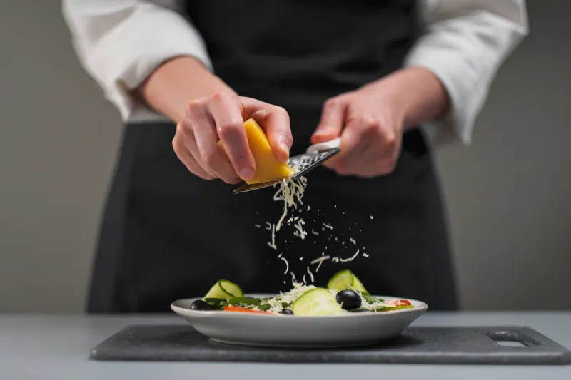 a female chef in a white uniform and a black apron in the restaurant kitchen cooking a salad of vegetables the cook rubs the parmesan cheese on a small grater
