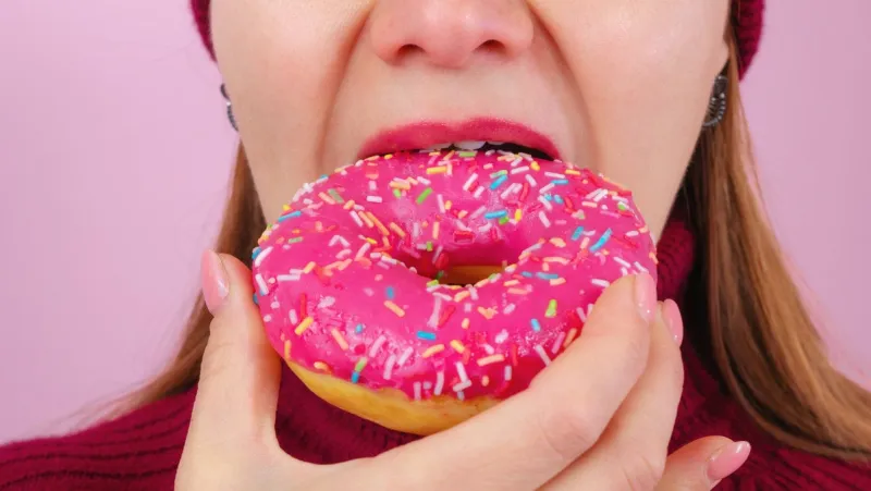 beautiful adult female in sweater and hat biting delicious donut against pink background close up