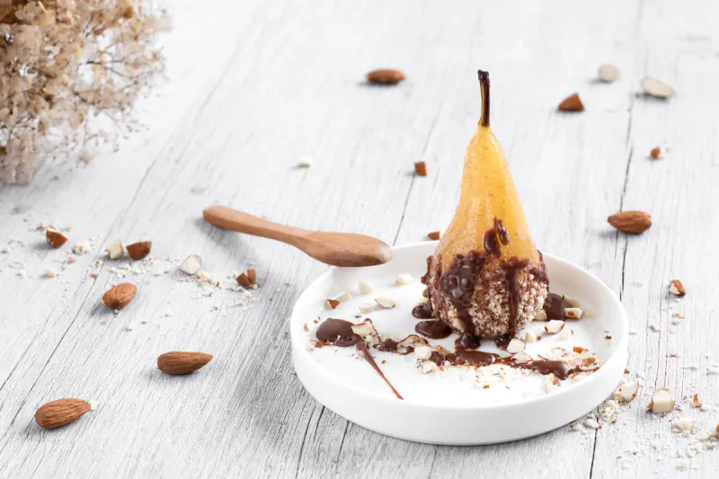 pear in chocolate and almonds on a white plate, on a wooden whit