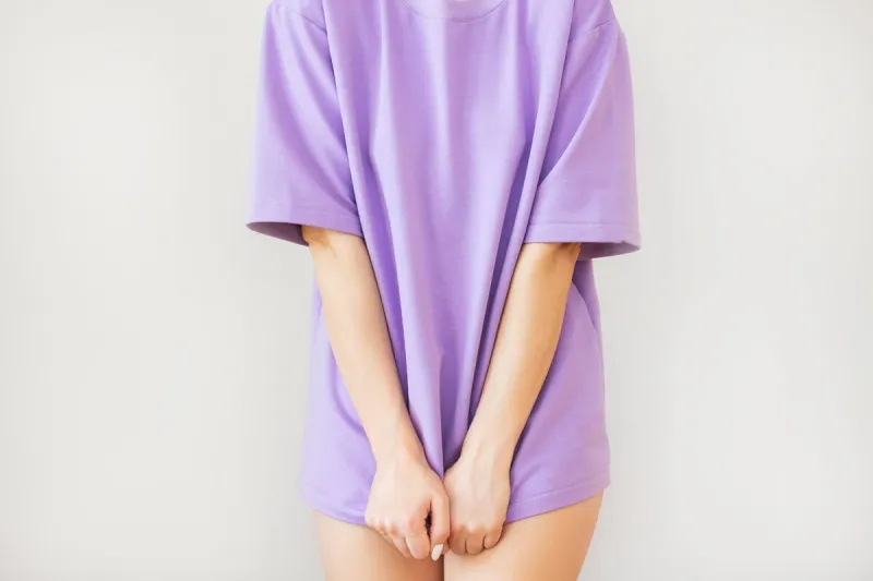 close-up of a young woman standing in a purple t-shirt and covering her crotch with her hands, isolated on a beige background suffers from urinary incontinence, cystitis, shy gynecological problems the concept of women's health