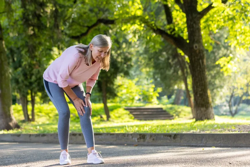 senior gray-haired woman walks and does sports in the park, stands bent over in pain, holds hands on knee