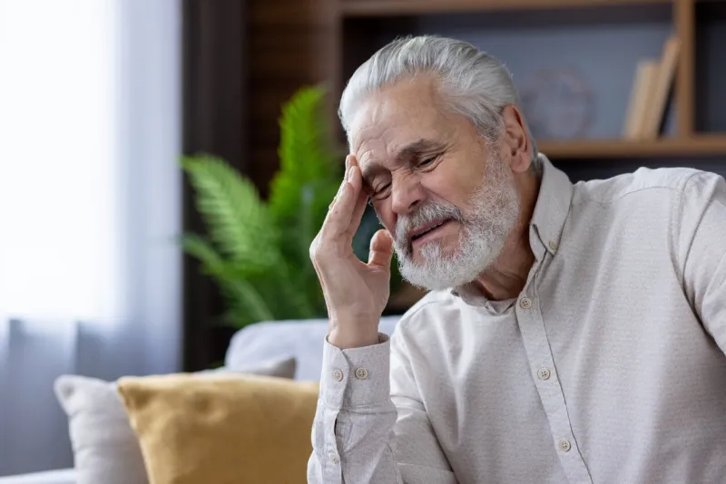 close-up photo of a tired and sick old gray-haired man sitting at home on the sofa and holding his head with his hand, closed his eyes and is in pain