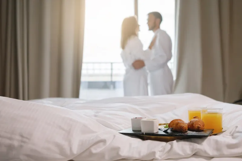 breakfast tray is on a white bed a couple in bathrobes are drinking coffee by the window in the background selective focus