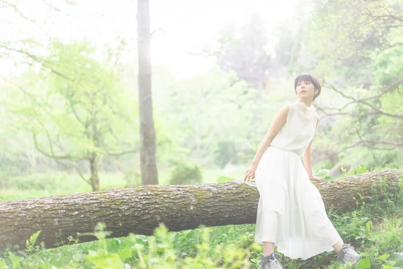 a young asian woman is sitting on a fallen tree sunlight filtering through treesback light