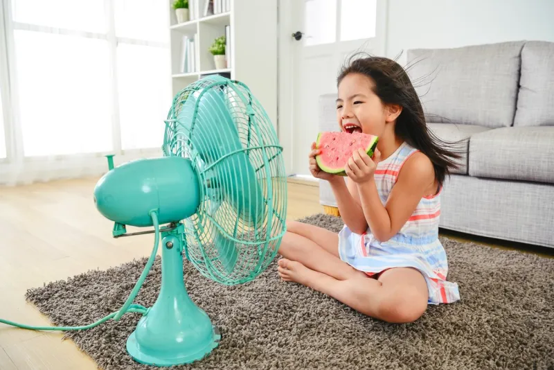 child eating watermelon with two hands sitting in front of the electric fan taking a big bite delightfully