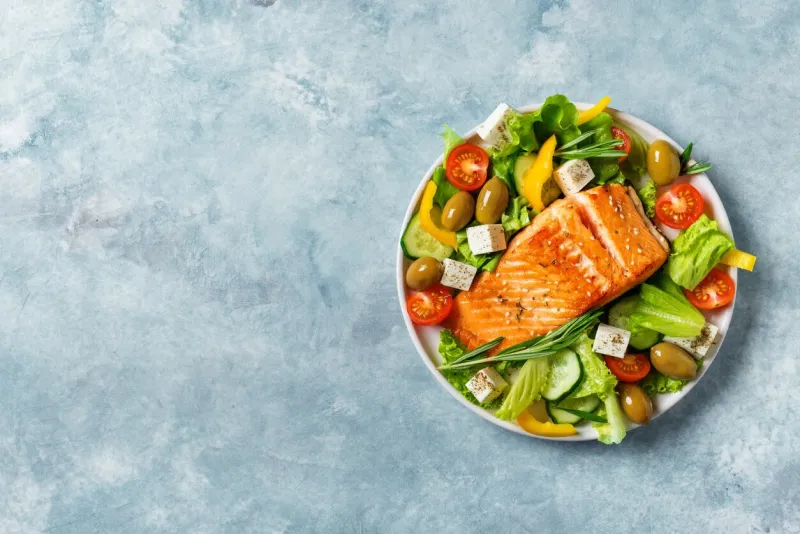 grilled salmon fish fillet and fresh vegetable salad with tomato, olives, lettuce, feta cheese mediterranean food - greek salad and roasted salmon on blue background, copy space