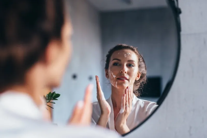 woman washes in front of the mirror, applying foam to her face