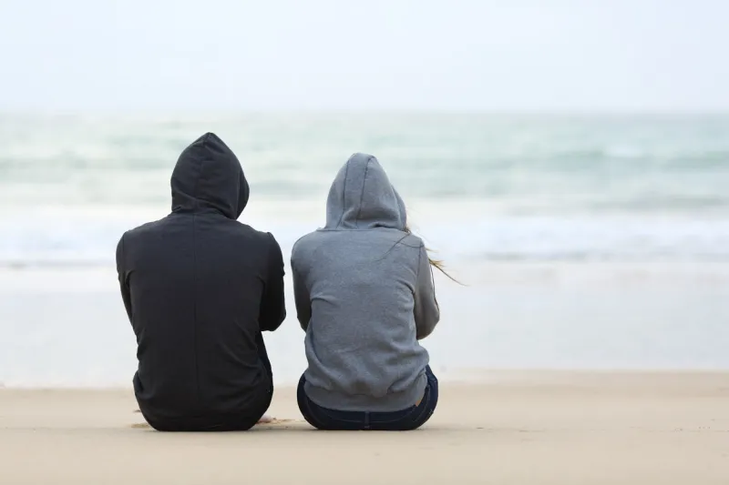 back view of two sad teenagers sitting on the sand of the beach and looking at horizon in a bad weather day