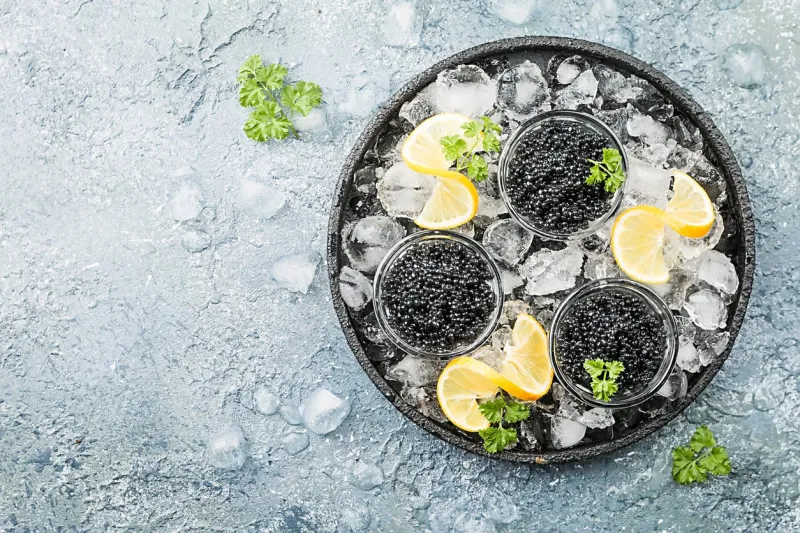 black caviar in a glass bowl on ice with lemon close up over gray background, top view