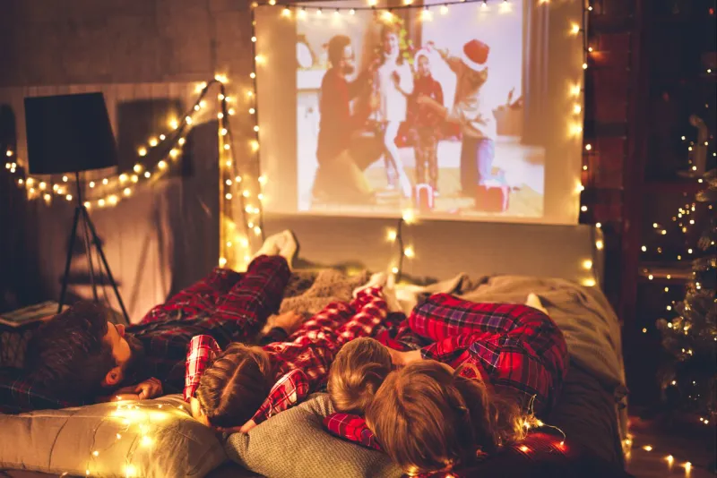 happy family in checkered pajamas  mother father and children watching projector, tv, movies with popcorn in christmas holiday evening  at home