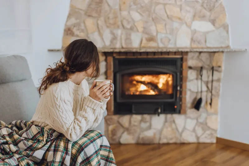 young woman sitting at home by the fireplace with a hot tea or coffee mug and warming her hands, she is wearing white woollen sweater cold houses in europe concept during energy and gas crisis