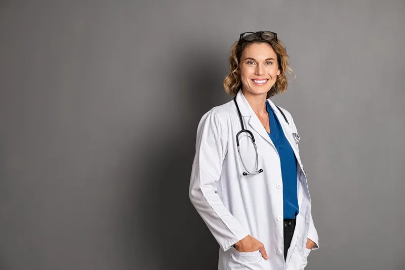 confident mature doctor in labcoat with stethoscope looking at camera smiling general pratictioner with hands in pockets standing isolated on grey background happy reliable physician isolated on gray wall with copy space