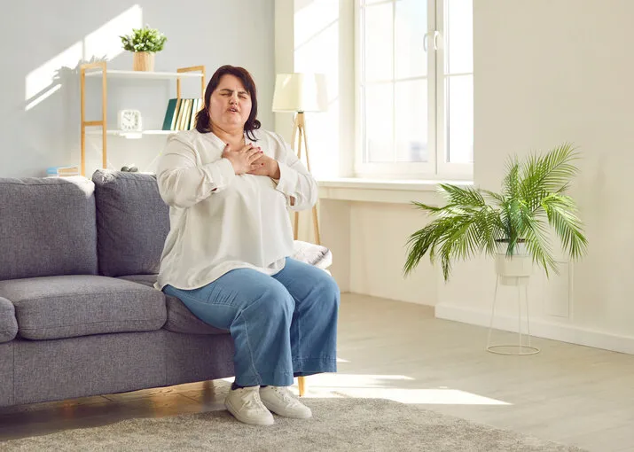 portrait of a young plump overweight fat woman sitting on the couch in the living room at home putting a hand on her chest suffering from heart problems obesity problems and healthcare concept