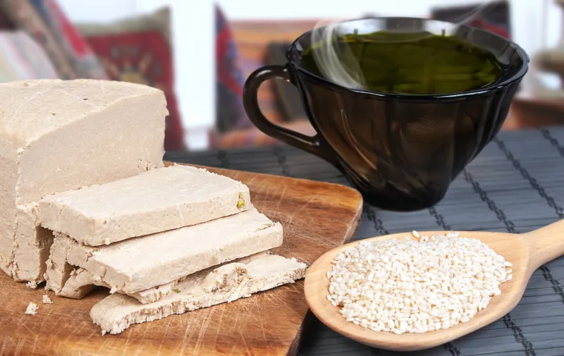sliced tahini halva, wooden spoon with sesame seeds and dark glass teacup close up