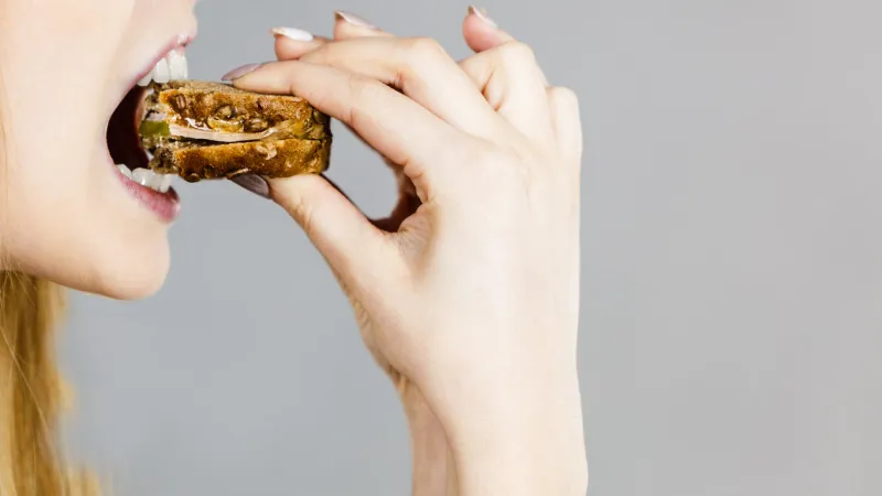 young woman eating sandwich, taking bite with wide open mouth food, calories, dieting concept studio shot on grey background, profile view