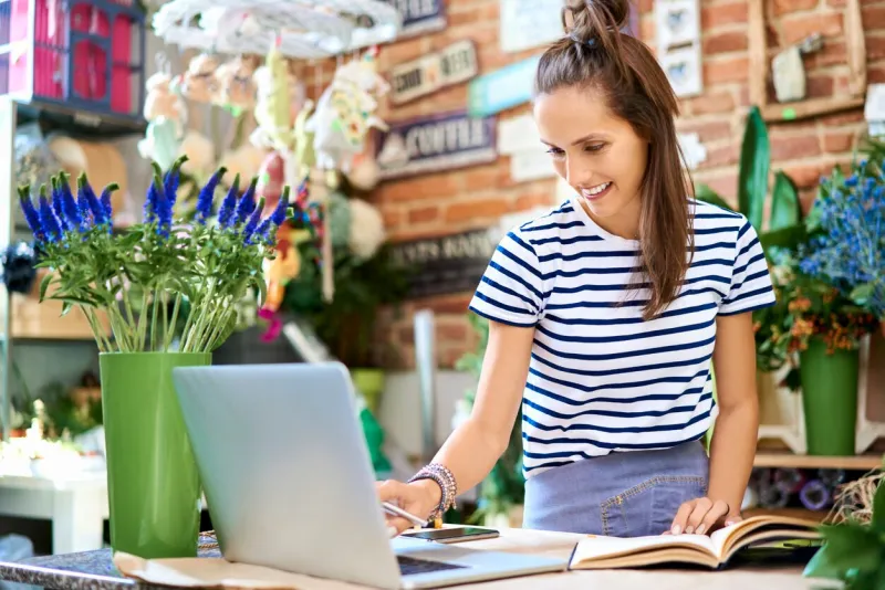 cheerful florist looking at laptop and writing down client orders in notebook